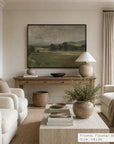 a framed landscape art print hangs on a beige wall in a styled living room. the print depicts a muted green and brown field scene with trees and hills. __lifestyleimage