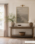 forgotten road in a gold floater frame under a brass picture light in a paneled entryway, above a wooden console with a vase of greenery, a ceramic lamp, and a woven basket. shown in floater gold, 30 x 40.