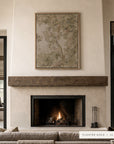 A framed art print with a floating gold border hangs above a rustic wood mantel and fireplace in a modern, neutral-toned living room. The print features a soft, painterly floral design in muted beige, sage green, and gray tones.