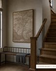 A large framed wall art print featuring a sepia-toned tree landscape hangs on a beige wall in a staircase landing. A dark wood bench with a gray throw sits below the artwork, and a wooden staircase with black metal railings is to the right.
