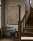 A large framed wall art print with a mottled beige and taupe floral pattern hangs on a warm beige wall in a styled entryway. To the left is a dark wood bench with a gray throw blanket, and to the right is a wooden staircase with black metal balusters.