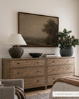 a floating walnut dresser in a rustic, warm wood finish, styled in a bedroom setting with a lamp, framed landscape painting, and a potted plant.