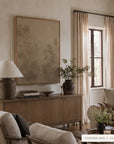 A large framed wall art print with a muted abstract landscape design hangs on a beige wall in a styled living room. The print has a light wood frame and is displayed above a wooden sideboard with a lamp, books, and a vase.