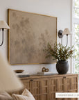 Large framed wall art with a muted beige and taupe abstract landscape design, bordered in gold, hanging in a styled room above a wooden credenza with two black wall sconces.