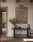 A framed landscape art print in a warm beige and brown tone hangs on a cream-colored wall above a rustic dark wood console table. The scene is styled with neutral ceramic vases, books, and dried botanicals, framed by rustic wooden doorposts. __lifestyleImage