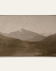 white mountains art print in a gold floater frame. a pale mountain peak rises over dark wooded hills and a still lake in warm sepia tones, shown against a plain off-white wall.