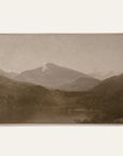 white mountains art print in a light oak floater frame. a pale mountain peak rises over dark wooded hills and a still lake in warm sepia tones, shown against a plain off-white wall.
