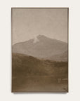 white mountains vertical art print in a gold floater frame. a pale mountain peak rises over dark wooded hills and a still lake in warm sepia tones, shown against a plain off-white wall.