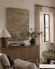 A framed landscape art print in a warm wood frame hangs on a beige wall above a wooden credenza in a styled living room. The print depicts a serene forest scene in muted earthy tones.