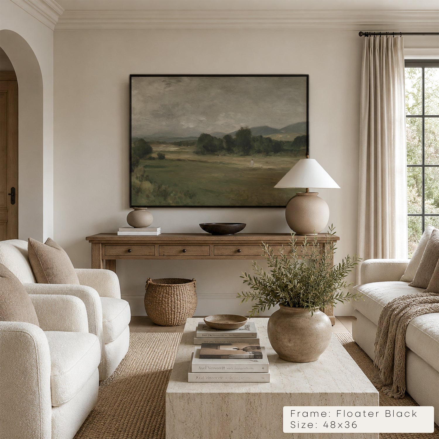A framed landscape art print hangs on a beige wall in a styled living room. The print depicts a muted green and brown field scene with trees and hills. __lifestyleImage