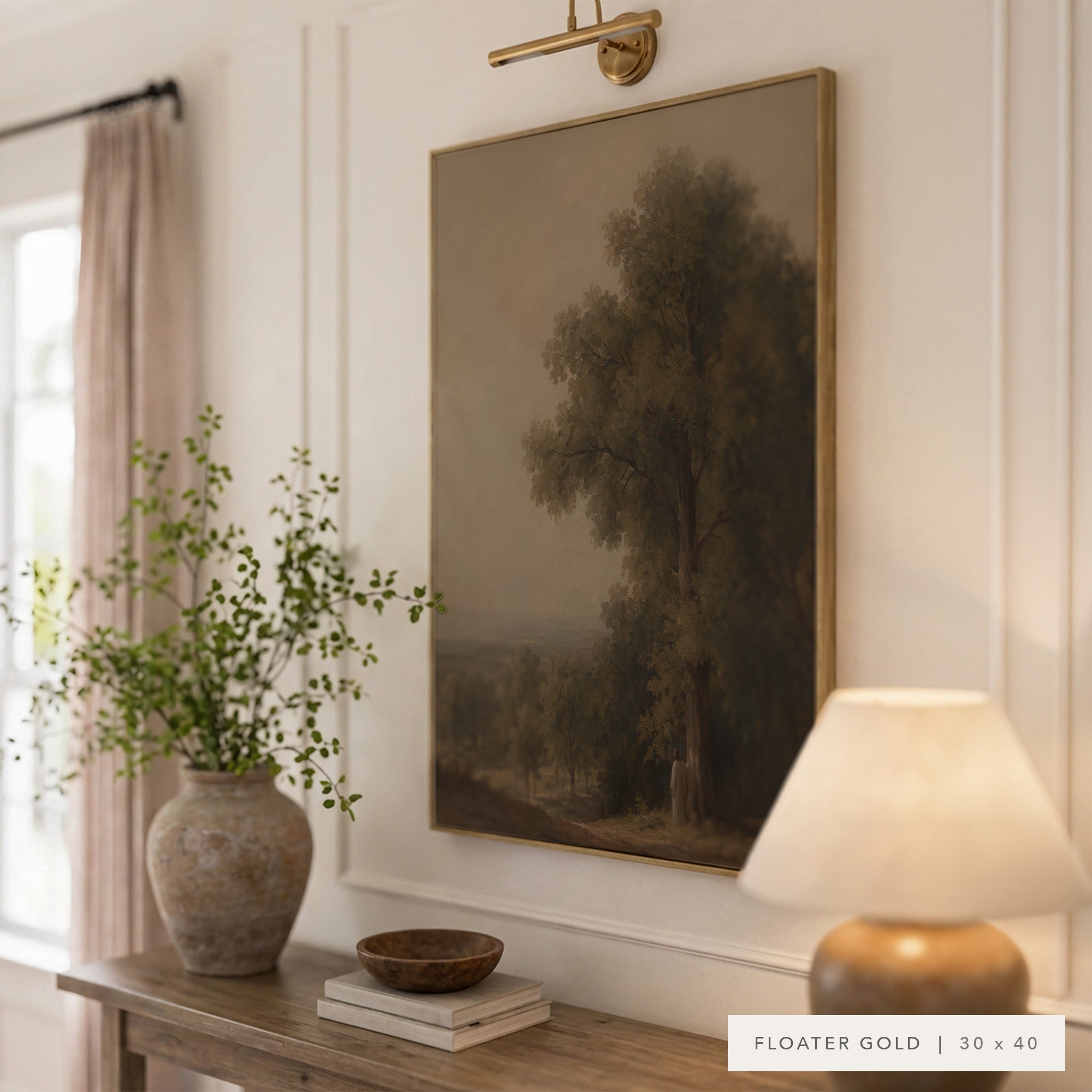A framed landscape art print featuring a tree in a muted forest scene, hung on a white paneled wall above a console table with a vase, bowl, and lamp. The frame is labeled as Floater Gold.