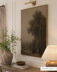A framed landscape art print featuring a tree in a muted forest scene, hung on a white paneled wall above a console table with a vase, bowl, and lamp. The frame is labeled as Floater Gold.