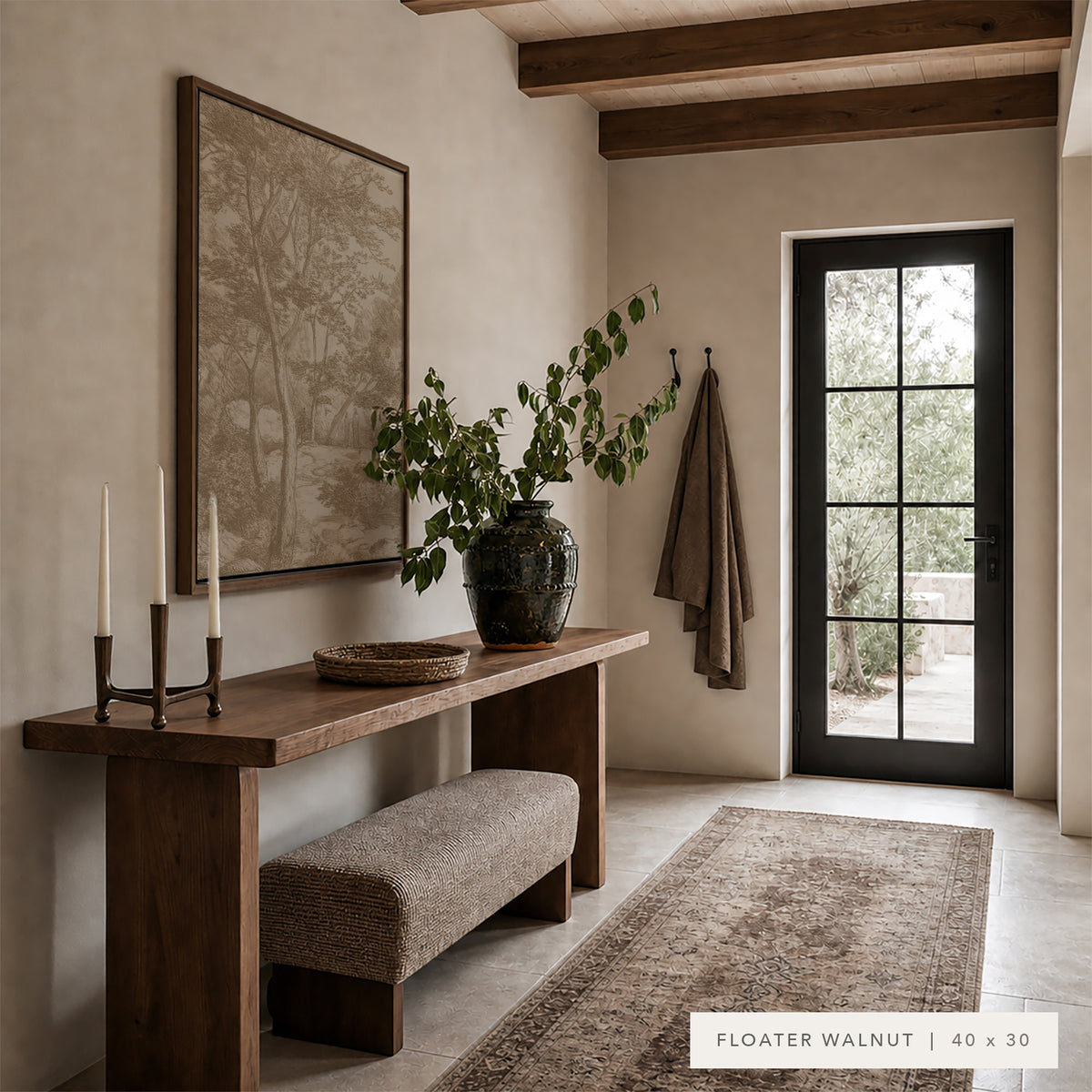 A floating walnut console table in a warm, rustic entryway. The table is styled with a large framed art print, a dark ceramic vase with green branches, and white taper candles.