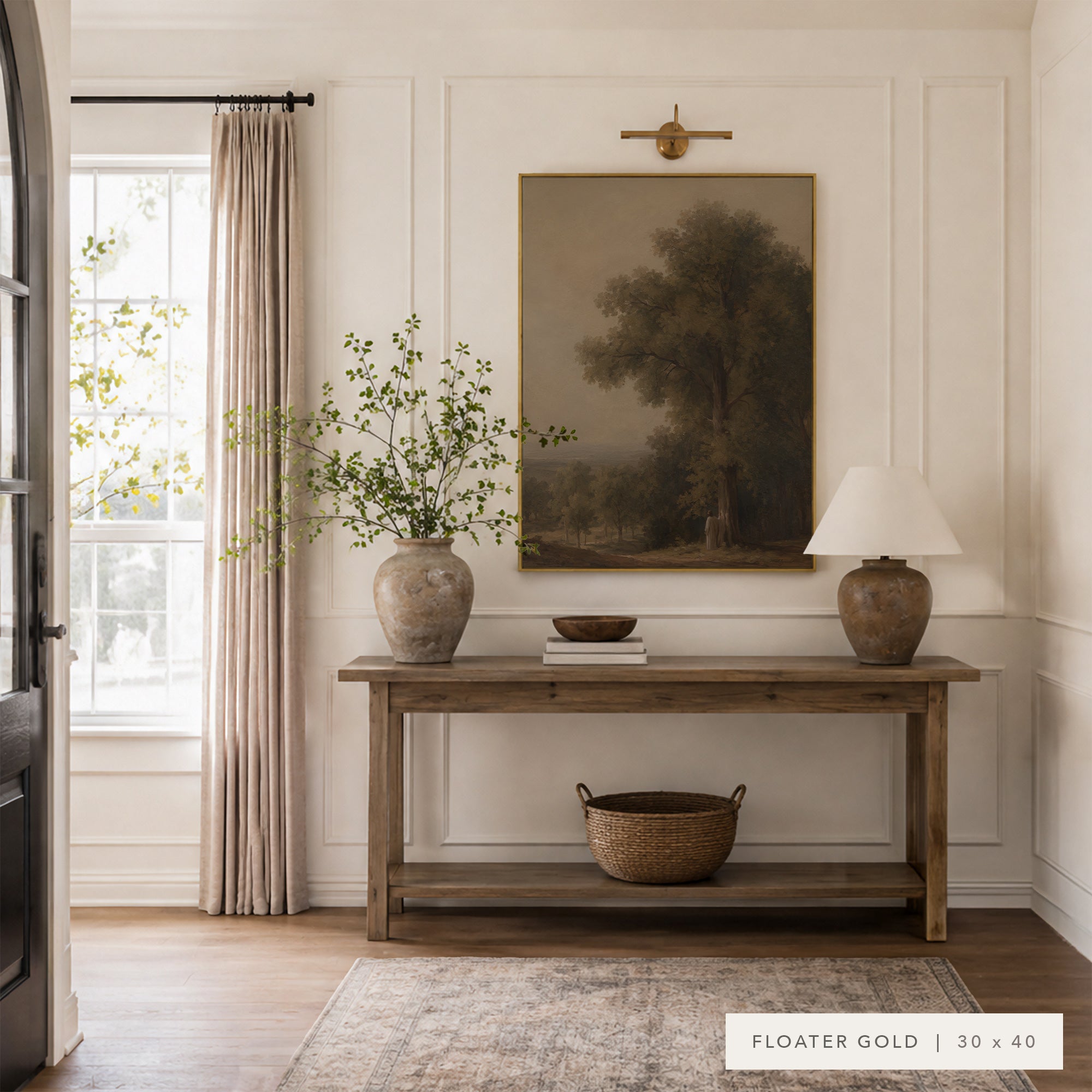 A framed landscape art print in a warm muted gold tone hangs on a white paneled wall above a wooden console table in a styled room. The scene includes a window with beige curtains, a vase with leafy branches, a table lamp, and a woven basket on the table. __lifestyleImage