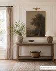 A framed landscape art print in a warm muted gold tone hangs on a white paneled wall above a wooden console table in a styled room. The scene includes a window with beige curtains, a vase with leafy branches, a table lamp, and a woven basket on the table. __lifestyleImage