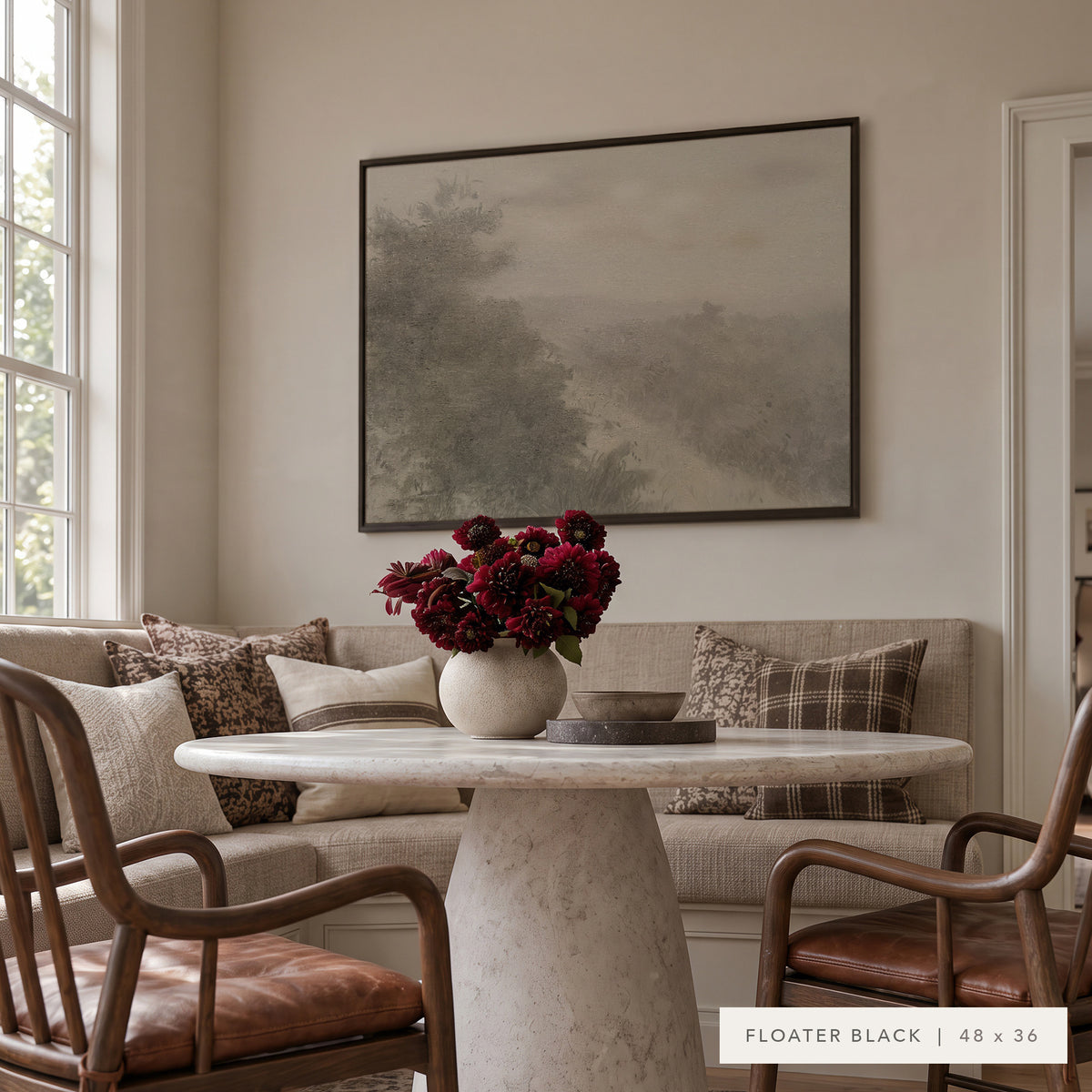 Forgotten Road in a black floater frame above a sunlit breakfast nook with a linen banquette, a round marble pedestal table, a vase of deep red flowers, and wooden armchairs with leather seats. Shown in Floater Black, 48 x 36. __lifestyleImage