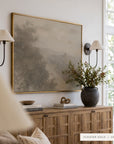 forgotten road in a gold floater frame between two black wall sconces with pleated shades, above a wooden sideboard styled with a dark vase of branches, books, and a small bowl. shown in floater gold, 48 x 36.