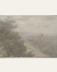 forgotten road art print in a light oak floater frame. a foggy road fades past a dark stand of trees in soft grays and warm stone tones, shown against a plain off-white wall. 