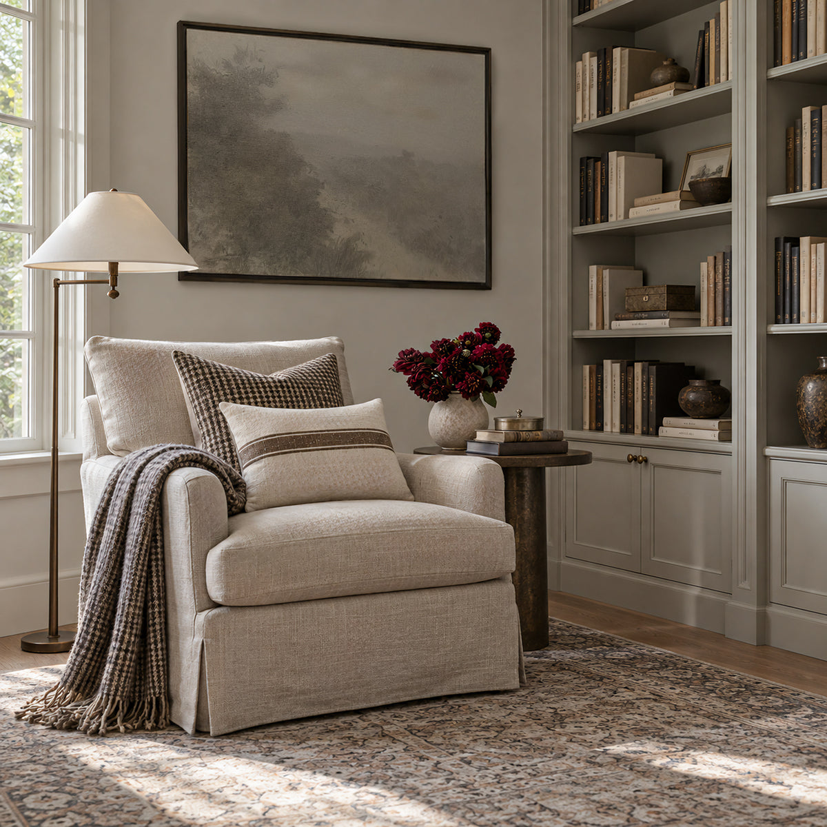 Forgotten Road in a dark frame above a cream slipcovered armchair with a plaid throw, beside a floor lamp, a side table with red flowers, and built-in bookshelves.