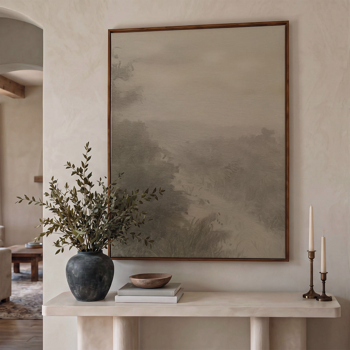 Forgotten Road in a dark frame above a cream plaster console with olive branches in a dark vase, a wooden bowl on books, and brass candlesticks.