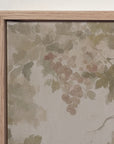 close-up of a framed wall art piece showing a painted grapevine with muted green leaves and blush pink grapes on a warm beige background, set in a light natural wood frame against a white wall.