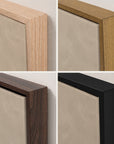 four close-up views of a square framed art print or canvas, showing different wood frame finishes: light natural wood, medium brown wood, dark walnut wood, and matte black. the background is a textured light beige wall.