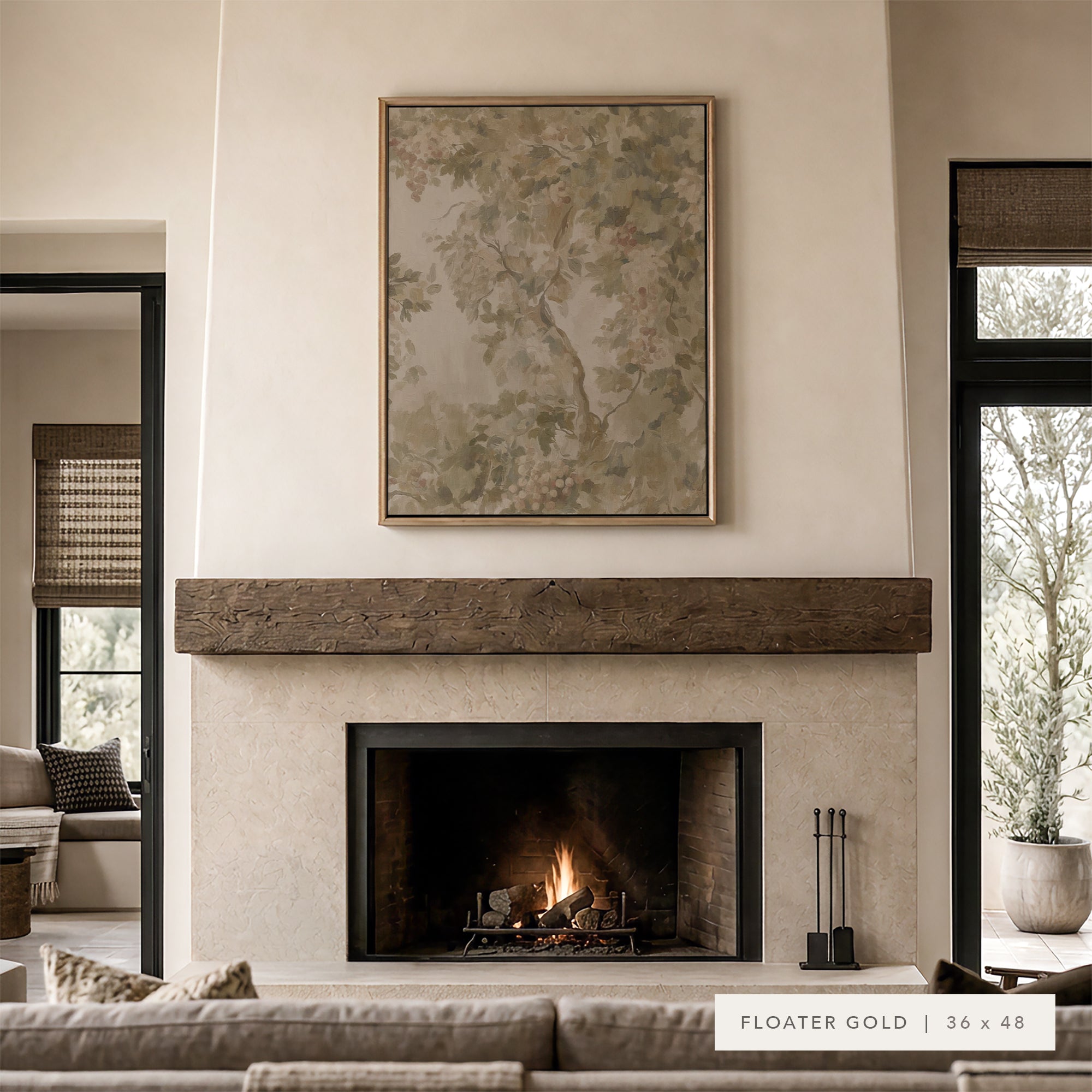 A framed art print with a floating gold border hangs above a rustic wood mantel and fireplace in a modern, neutral-toned living room. The print features a soft, painterly floral design in muted beige, sage green, and gray tones.