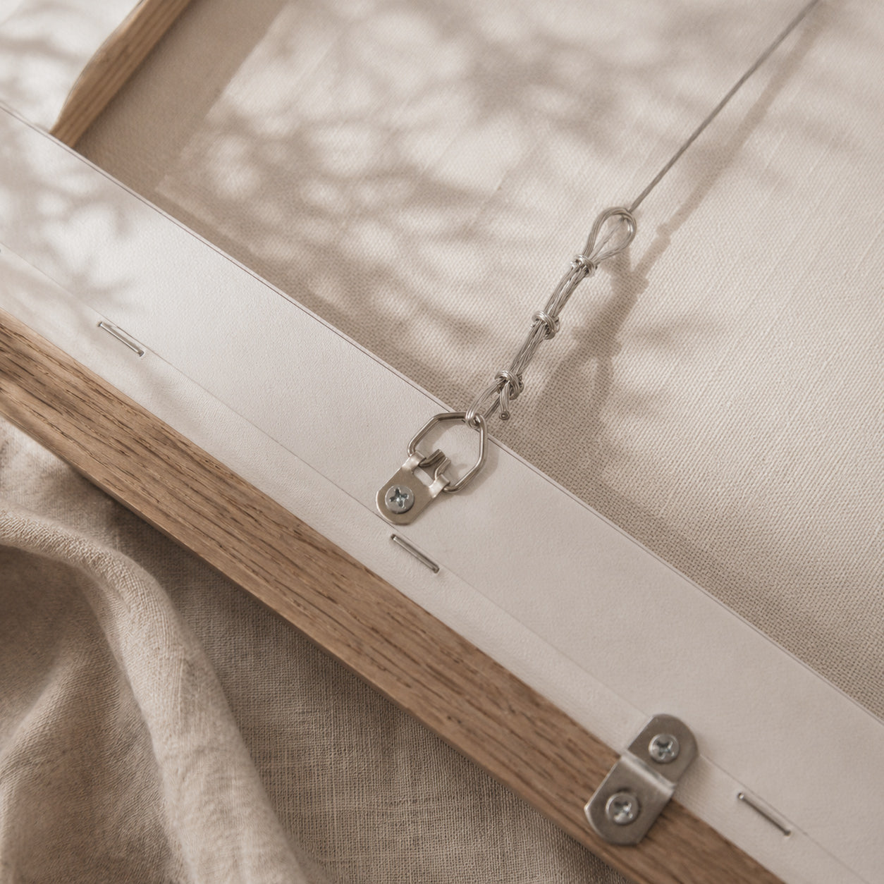 Close-up of a framed canvas or fabric artwork showing the wooden frame, metal hanging hardware, and textured off-white fabric with soft leaf shadows.