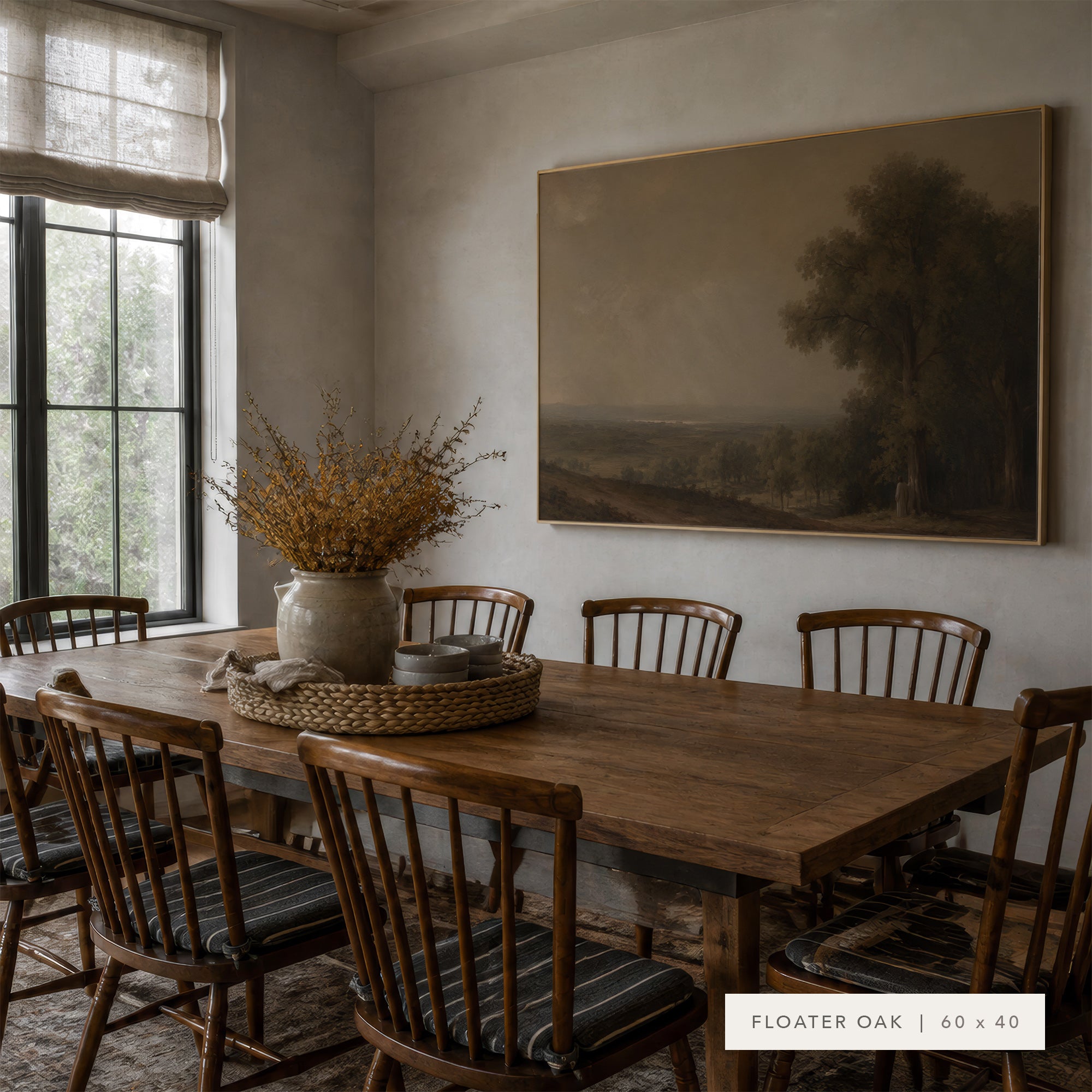 A large framed landscape painting in a warm wood frame hangs on a beige wall in a dining room. The room features a wooden dining table and matching wooden chairs with striped cushions, decorated with a ceramic vase of dried branches on a woven tray.