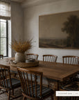 A large framed landscape painting in a warm wood frame hangs on a beige wall in a dining room. The room features a wooden dining table and matching wooden chairs with striped cushions, decorated with a ceramic vase of dried branches on a woven tray.