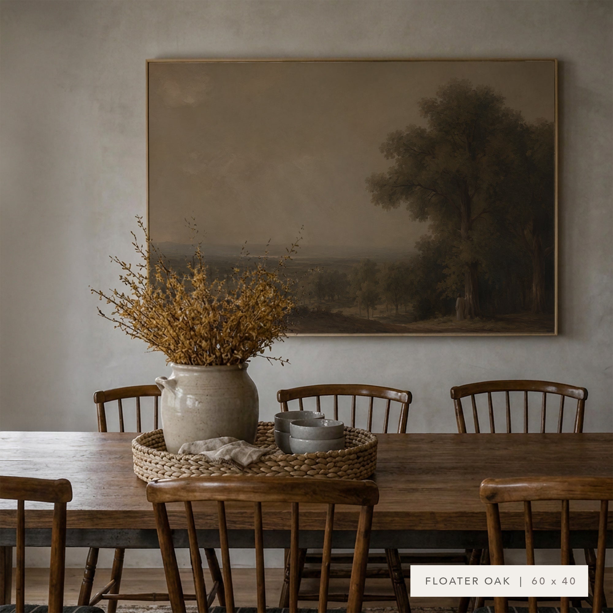 A large framed landscape art print in a warm brown tone hangs on a textured plaster wall above a wooden dining table with chairs and a vase of dried branches. __lifestyleImage