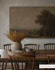 A large framed landscape art print in a warm brown tone hangs on a textured plaster wall above a wooden dining table with chairs and a vase of dried branches. __lifestyleImage