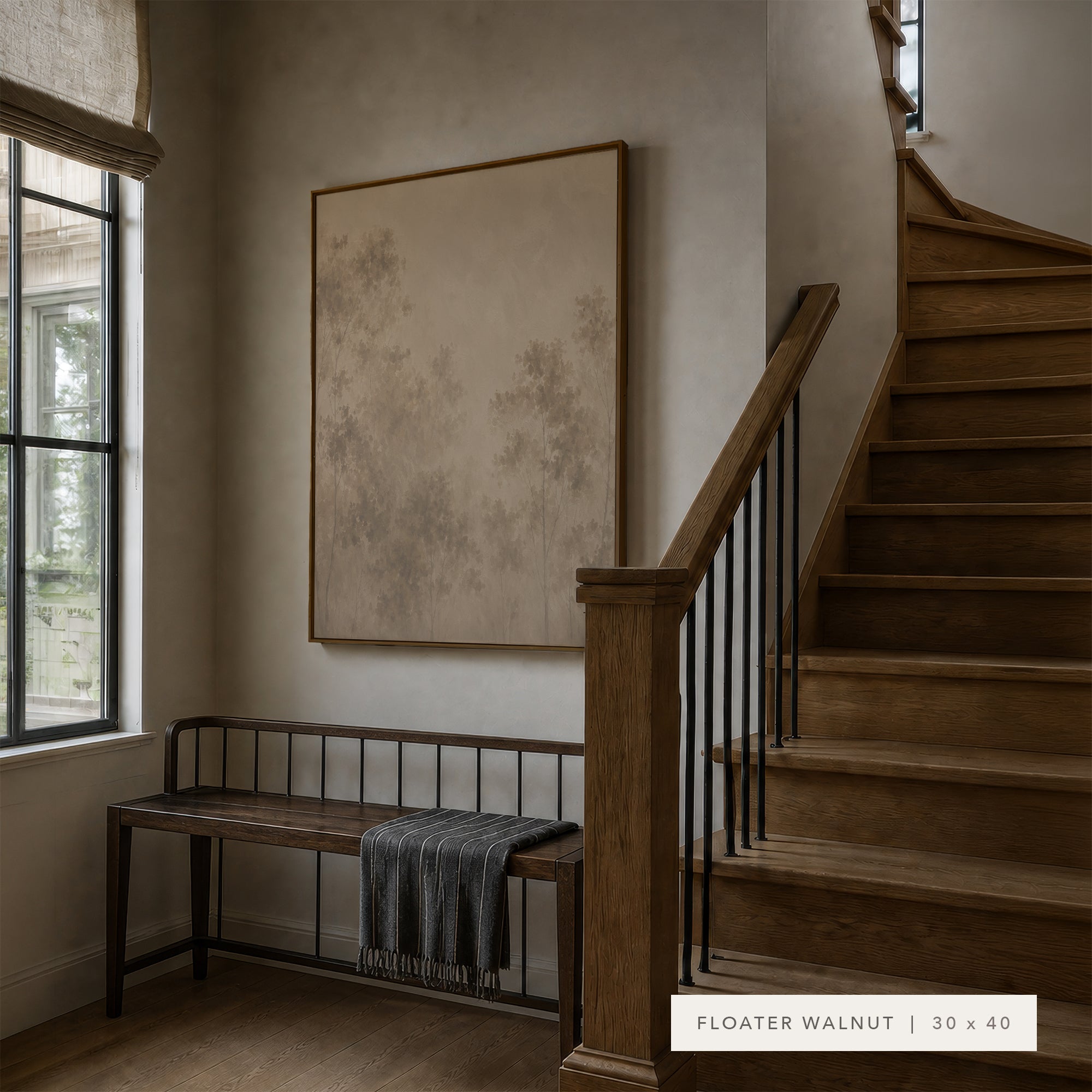 A large framed wall art print with a mottled beige and taupe floral pattern hangs on a warm beige wall in a styled entryway. To the left is a dark wood bench with a gray throw blanket, and to the right is a wooden staircase with black metal balusters.