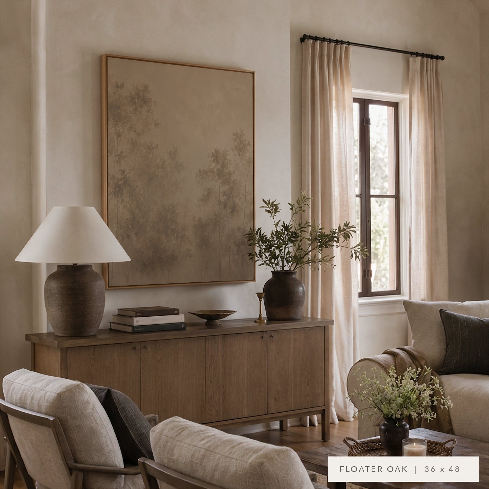 A large framed wall art print with a muted abstract landscape design hangs on a beige wall in a styled living room. The print has a light wood frame and is displayed above a wooden sideboard with a lamp, books, and a vase.