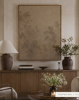 A large framed wall art print in a warm taupe and beige abstract landscape design, styled in a cozy living room setting above a wooden console table with a lamp, books, and a vase. __lifestyleImage