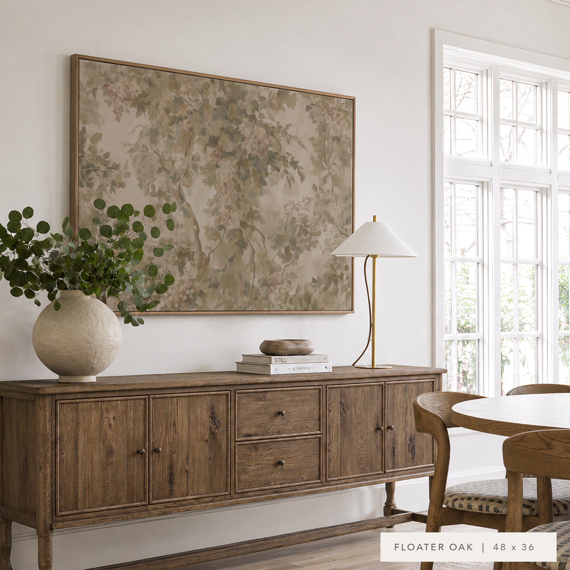 A large framed botanical art print in a warm beige and brown palette hangs on a white wall above a rustic wooden sideboard. The room is styled with a textured ceramic vase of green leaves, a brass lamp, books, and a wooden dining table with chairs near a bright window.