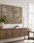 A large framed botanical art print in a warm beige and brown palette hangs on a white wall above a rustic wooden sideboard. The room is styled with a textured ceramic vase of green leaves, a brass lamp, books, and a wooden dining table with chairs near a bright window.