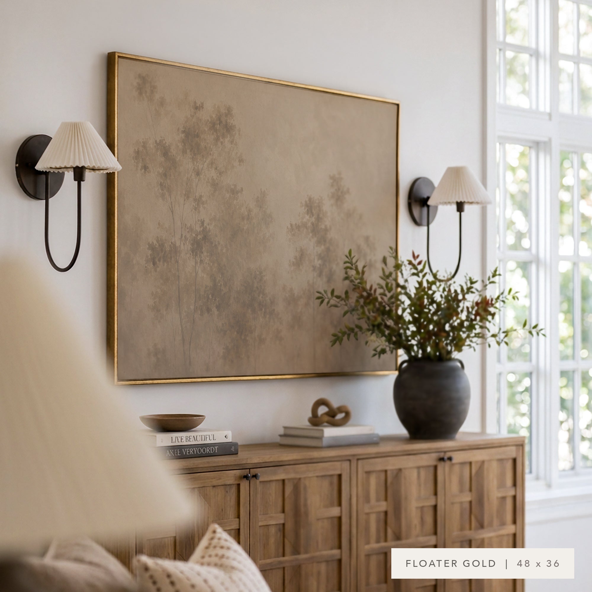 Large framed wall art with a muted beige and taupe abstract landscape design, bordered in gold, hanging in a styled room above a wooden credenza with two black wall sconces.