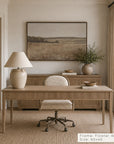 a framed landscape art print in a floater walnut frame hangs on a beige wall above a wooden console table in a styled home office. the print depicts a dry grass field under a soft sky. __lifestyleimage
