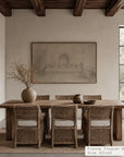 a large framed wall art print hangs above a rustic wooden dining table with woven chairs in a neutral dining room. the print depicts a soft sepia architectural scene. __lifestyleimage