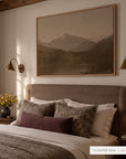white mountains in a light oak floater frame above an upholstered bed with floral and plum pillows, between two brass wall sconces, with yellow flowers on the nightstand. shown in floater oak, 60 x 40. __lifestyleimage