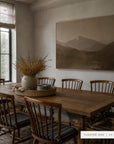 white mountains in a light oak floater frame above a long wooden farmhouse table with spindle-back chairs, a woven tray, and a crock of dried golden branches. shown in floater oak, 60 x 40.