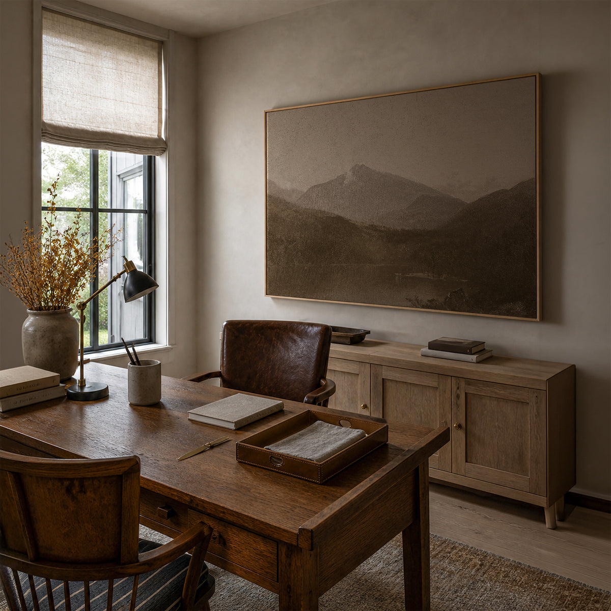 White Mountains in a light wood frame in a home office, above a wooden sideboard, with a wooden desk, a brass desk lamp, a leather armchair, and a crock of dried golden branches by the window.