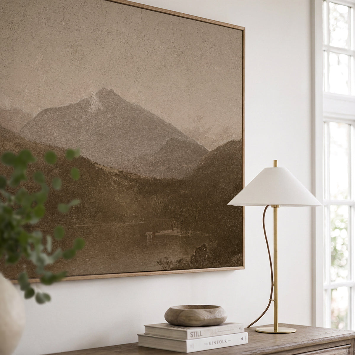 A close, angled view of White Mountains in a light wood frame beside tall windows, above a wooden console with a brass table lamp, a wooden bowl on books, and trailing greenery.