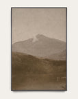 white mountains vertical art print in a black floater frame. a pale mountain peak rises over dark wooded hills and a still lake in warm sepia tones, shown against a plain off-white wall. 