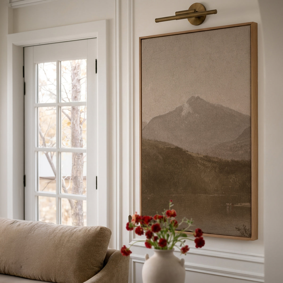 White Mountains in a light wood frame under a brass picture light, beside white French doors, above a camel sofa and a small vase of red flowers.