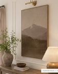 white mountains in a gold floater frame under a brass picture light, above a wooden console with a vase of greenery, a wooden bowl on books, and a glowing table lamp. shown in floater gold, 30 x 40.