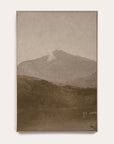 white mountains vertical art print in a light oak floater frame. a pale mountain peak rises over dark wooded hills and a still lake in warm sepia tones, shown against a plain off-white wall. 