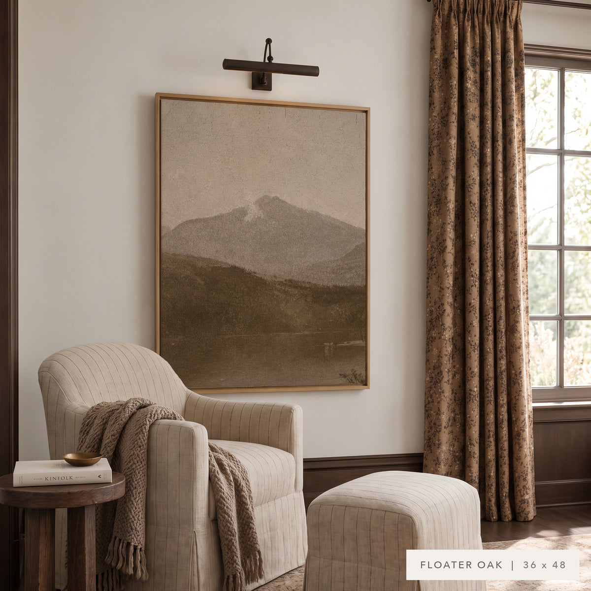 White Mountains in a light oak floater frame under a bronze picture light, above a cream striped armchair and ottoman with a knit throw, beside patterned drapes and a small wooden side table. Shown in Floater Oak, 36 x 48. __lifestyleImage