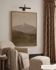 white mountains in a light oak floater frame under a bronze picture light, above a cream striped armchair and ottoman with a knit throw, beside patterned drapes and a small wooden side table. shown in floater oak, 36 x 48. __lifestyleimage